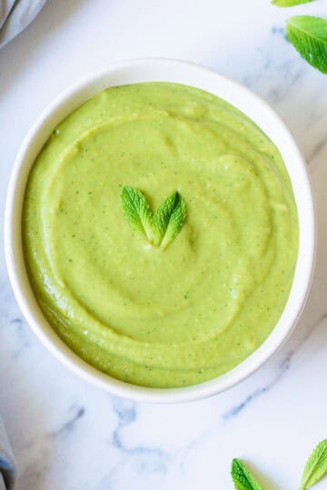 A white bowl filled pea and mint soup, garnished with two fresh mint leaves, sits on a marble surface with a few loose mint leaves and a light blue cloth nearby.
