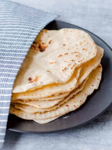 Homemade flour tortillas on black plate half covered with blue and white striped tea towel.