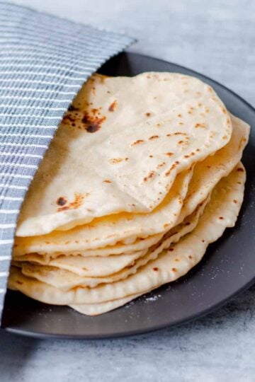 Homemade flour tortillas on black plate half covered with blue and white striped tea towel.