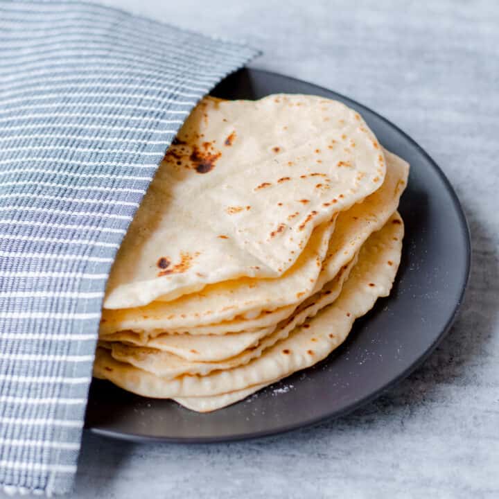 Homemade flour tortillas on black plate half covered with blue and white striped tea towel.