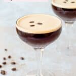 A coupe glass filled with a dark espresso martini, topped with three coffee beans. A few scattered coffee beans are on the light textured surface nearby, and a second glass is blurred in the background.
