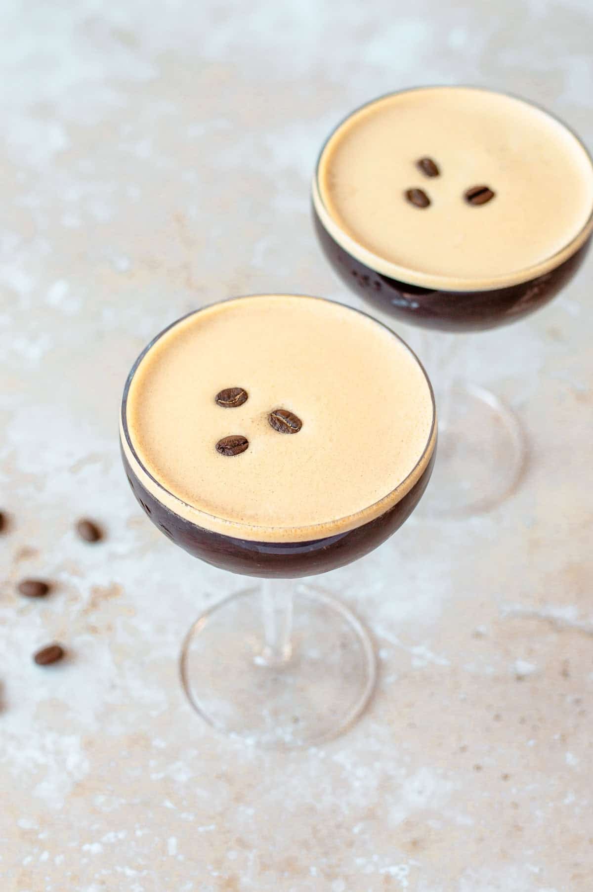 Two espresso martinis in coupe glasses, each topped with three coffee beans, are displayed on a light, textured surface with a few scattered coffee beans nearby.