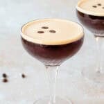 A coupe glass filled with a dark espresso martini, topped with three coffee beans. A few scattered coffee beans are on the light textured surface nearby, and a second glass is blurred in the background.