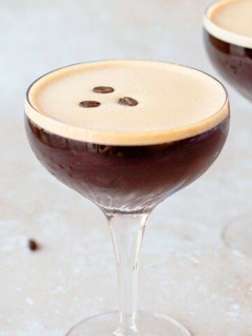 A coupe glass filled with a dark espresso martini, topped with three coffee beans. A few scattered coffee beans are on the light textured surface nearby, and a second glass is blurred in the background.