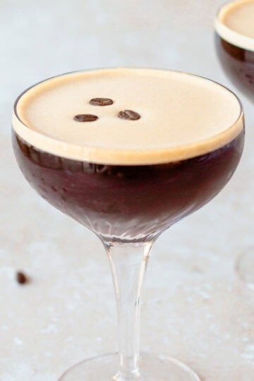 A coupe glass filled with a dark espresso martini, topped with three coffee beans. A few scattered coffee beans are on the light textured surface nearby, and a second glass is blurred in the background.