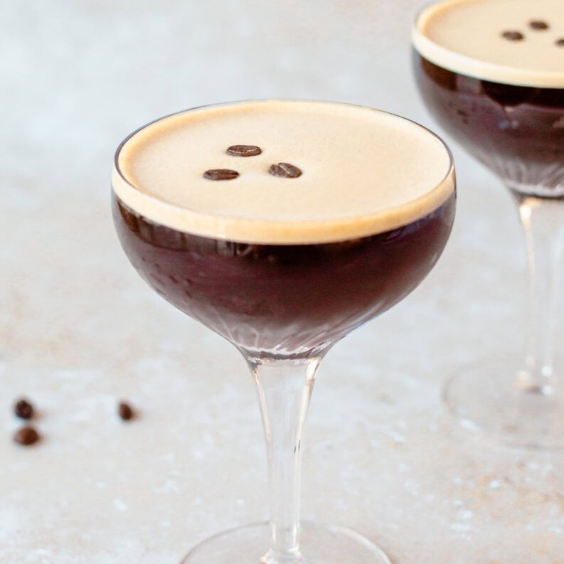 A coupe glass filled with a dark espresso martini, topped with three coffee beans. A few scattered coffee beans are on the light textured surface nearby, and a second glass is blurred in the background.