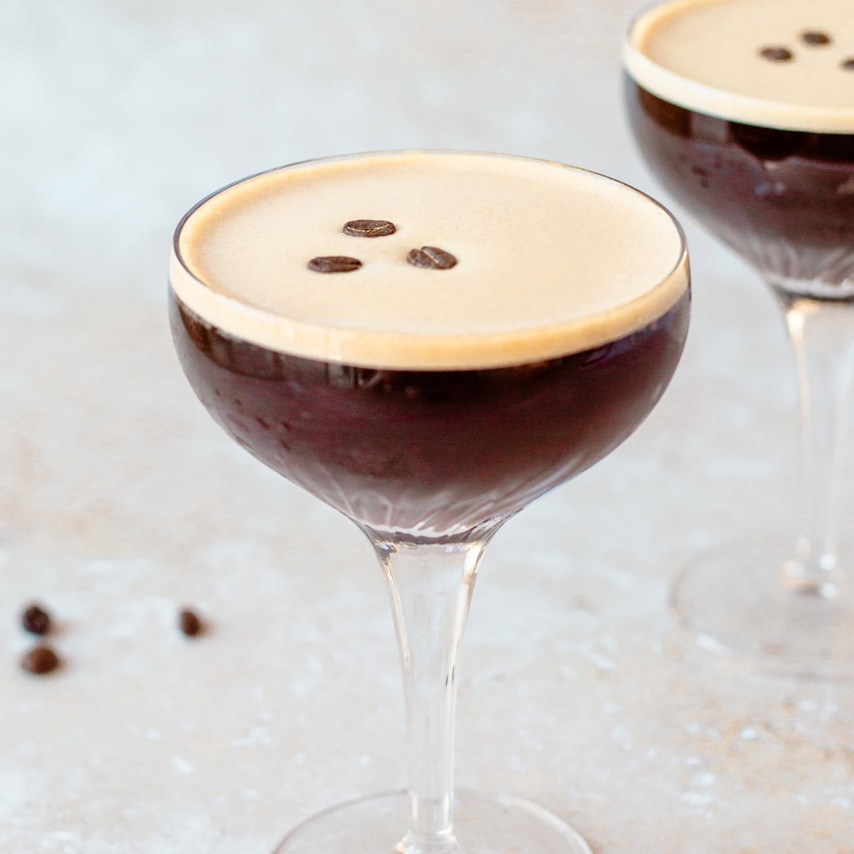 A coupe glass filled with a dark espresso martini, topped with three coffee beans. A few scattered coffee beans are on the light textured surface nearby, and a second glass is blurred in the background.