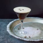 A martini glass filled with an Espresso Martini with 3 coffee beans on top. It's sitting on a metal tray on a dark background.
