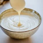 A white bechamel sauce in a pale sage colour bowl. A spoon is above the bowl with some bechamel sauce dripping off it.