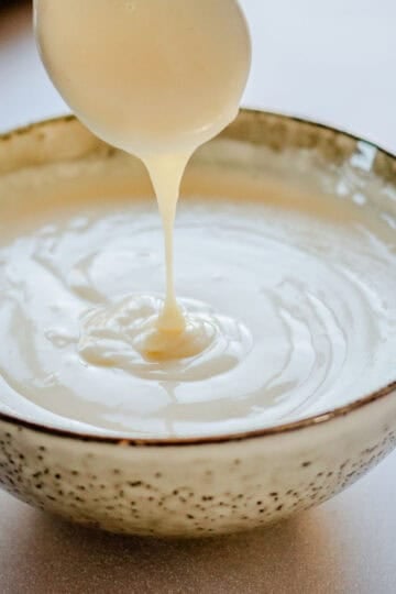 A white bechamel sauce in a pale sage colour bowl. A spoon is above the bowl with some bechamel sauce dripping off it.
