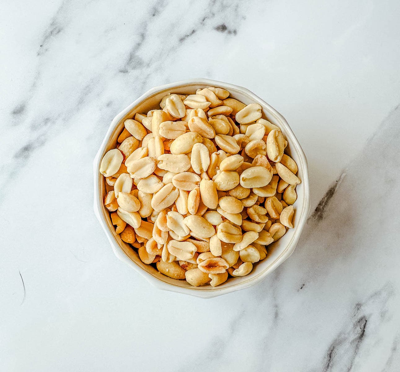A white bowl filled with shelled, lightly roasted peanuts sits on a white marble surface.