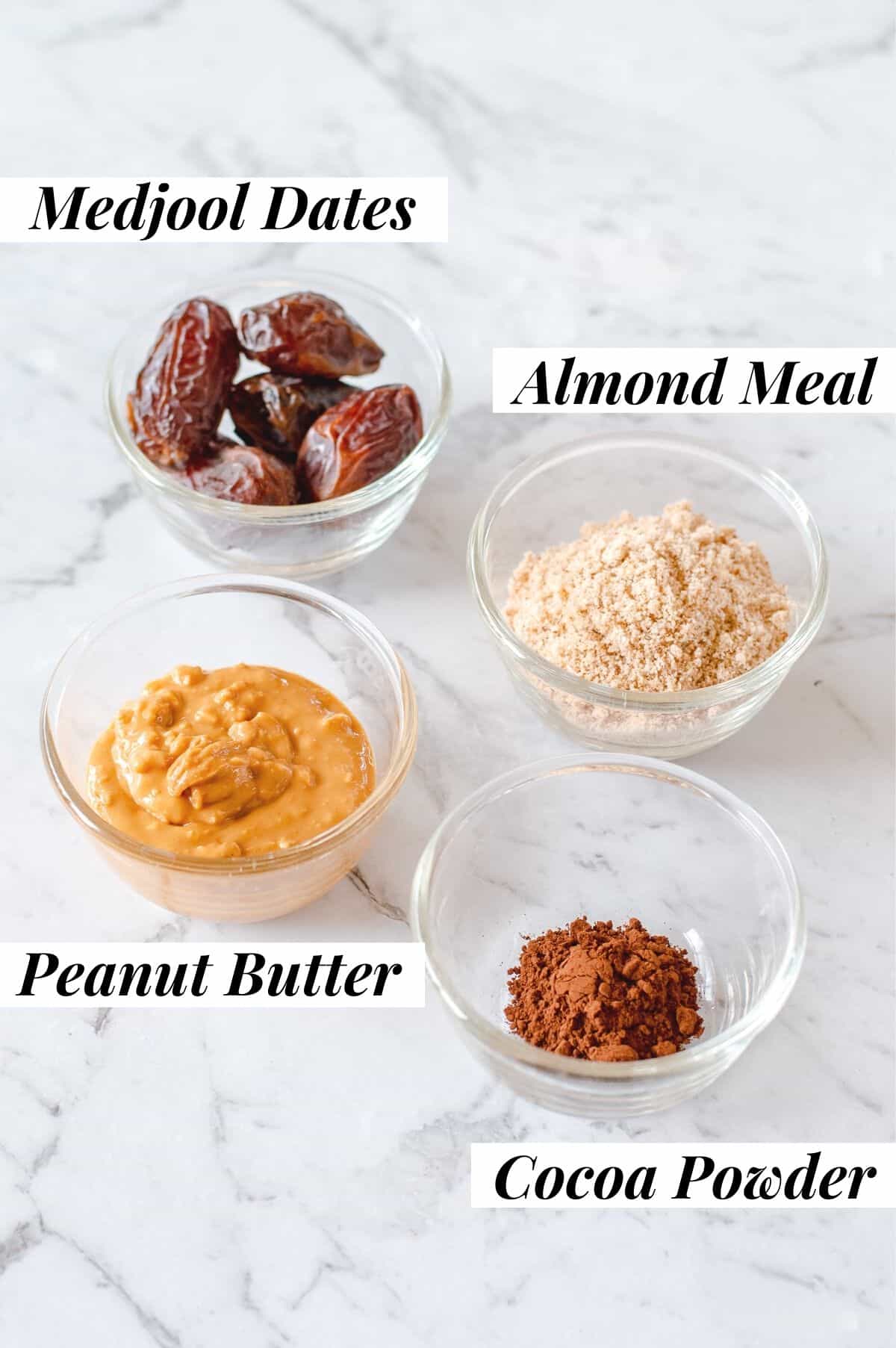 An image of dates, peanut butter, almond meal and cocoa powder in glass bowls on marble background.