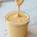 A spoon lifts creamy peanut butter from a glass jar, creating a smooth, thick drizzle; a few peanut pieces rest on the white marble surface nearby.