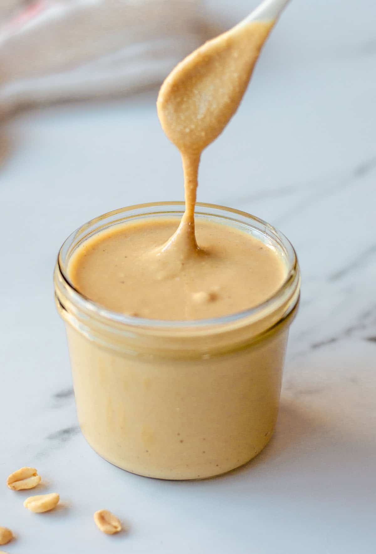 A spoon lifts creamy peanut butter from a glass jar, creating a smooth, thick drizzle; a few peanut pieces rest on the white marble surface nearby.