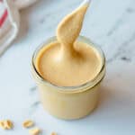 A spoon lifts creamy peanut butter from a glass jar on a white marble surface, with a few peanuts scattered nearby.