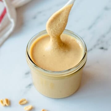 A spoon lifts creamy peanut butter from a glass jar on a white marble surface, with a few peanuts scattered nearby.