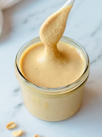 A spoon lifts creamy peanut butter from a glass jar on a white marble surface, with a few peanuts scattered nearby.