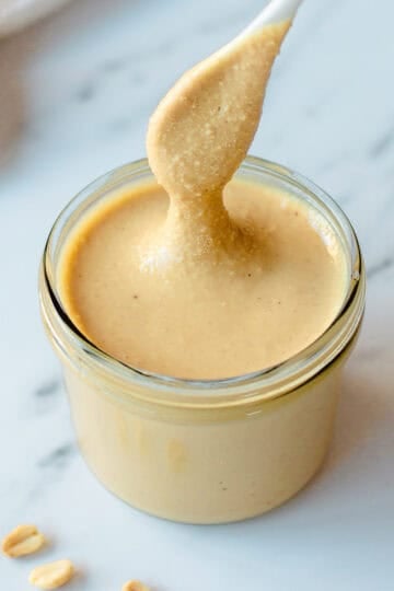 A spoon lifts creamy peanut butter from a glass jar on a white marble surface, with a few peanuts scattered nearby.
