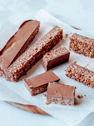 Seven chocolate-coated mars bar slices are arranged on baking parchment atop a round wooden board. The bars have a thick chocolate layer on top and a textured, grainy interior.