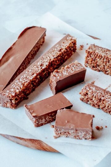 Seven chocolate-coated mars bar slices are arranged on baking parchment atop a round wooden board. The bars have a thick chocolate layer on top and a textured, grainy interior.
