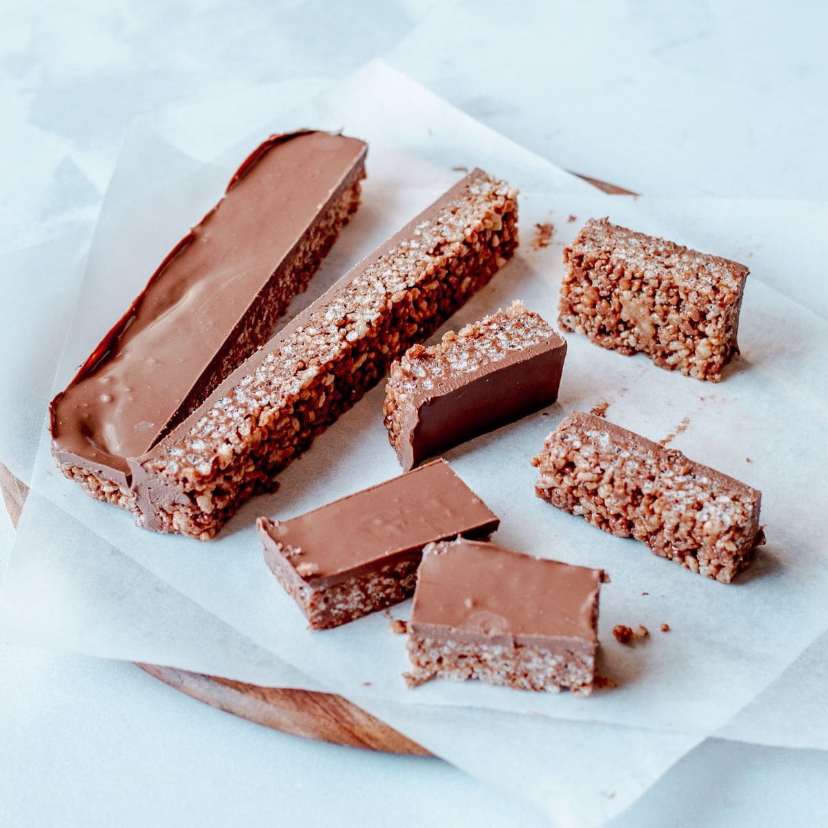 Seven chocolate-coated mars bar slices are arranged on baking parchment atop a round wooden board. The bars have a thick chocolate layer on top and a textured, grainy interior.