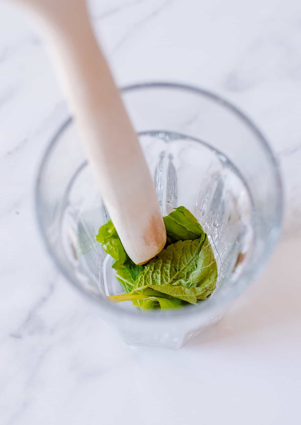 Mint leaves being muddled in a glass with a wooden spoon.