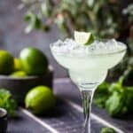 Lime Margarita in a tall glass with limes in the background.