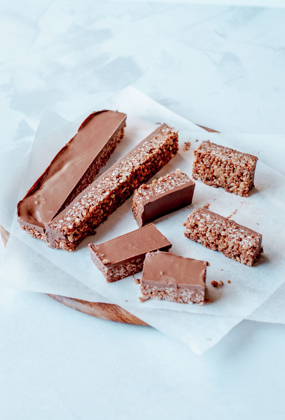 Seven chocolate-coated mars bar slices are arranged on baking parchment atop a round wooden board. The bars have a thick chocolate layer on top and a textured, grainy interior.