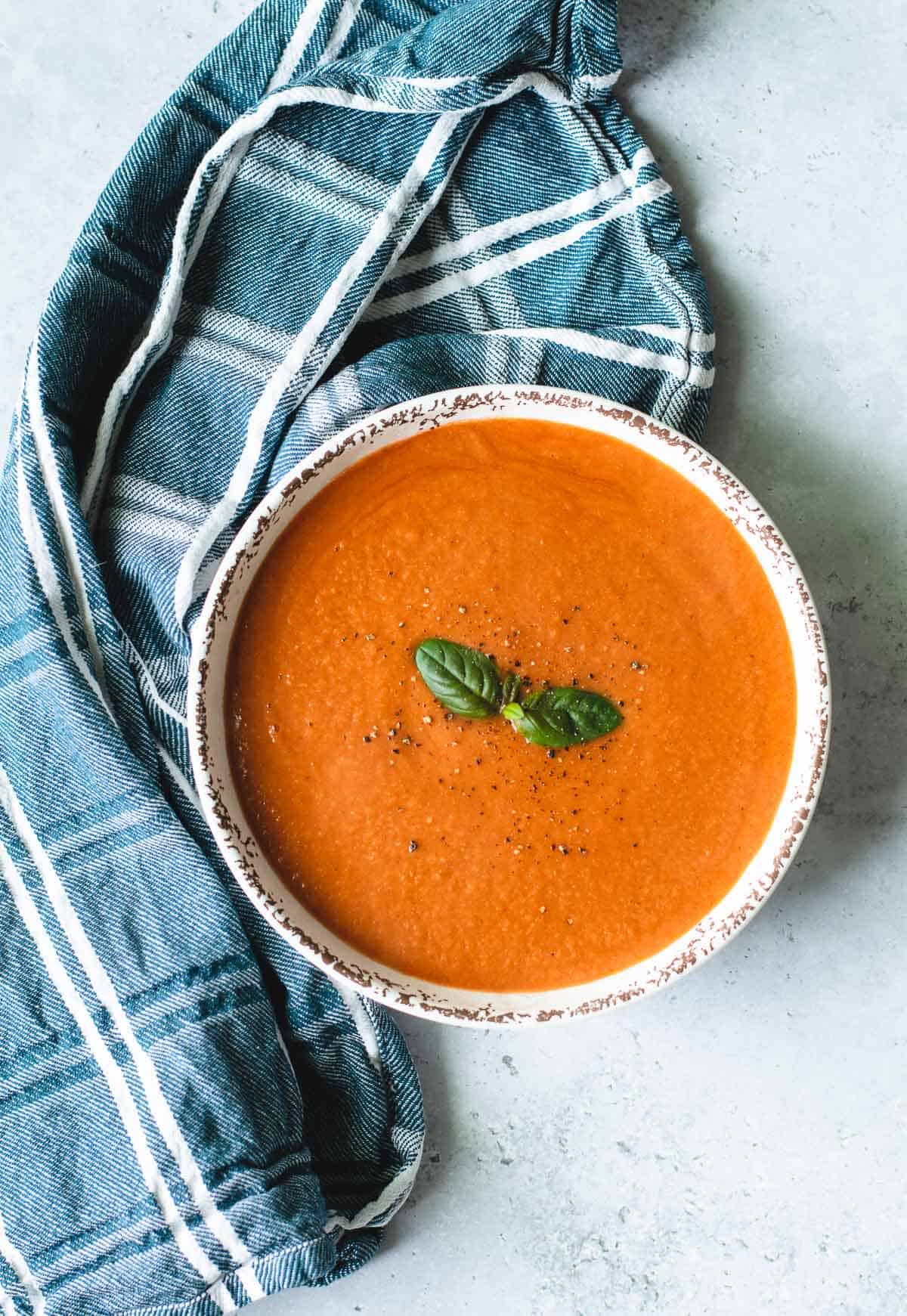 Tomato Soup in a white bowl sitting on a grey background with blue and white tea towel.