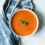 Tomato soup in a white bowl sitting on a grey background with blue and white tea towel.