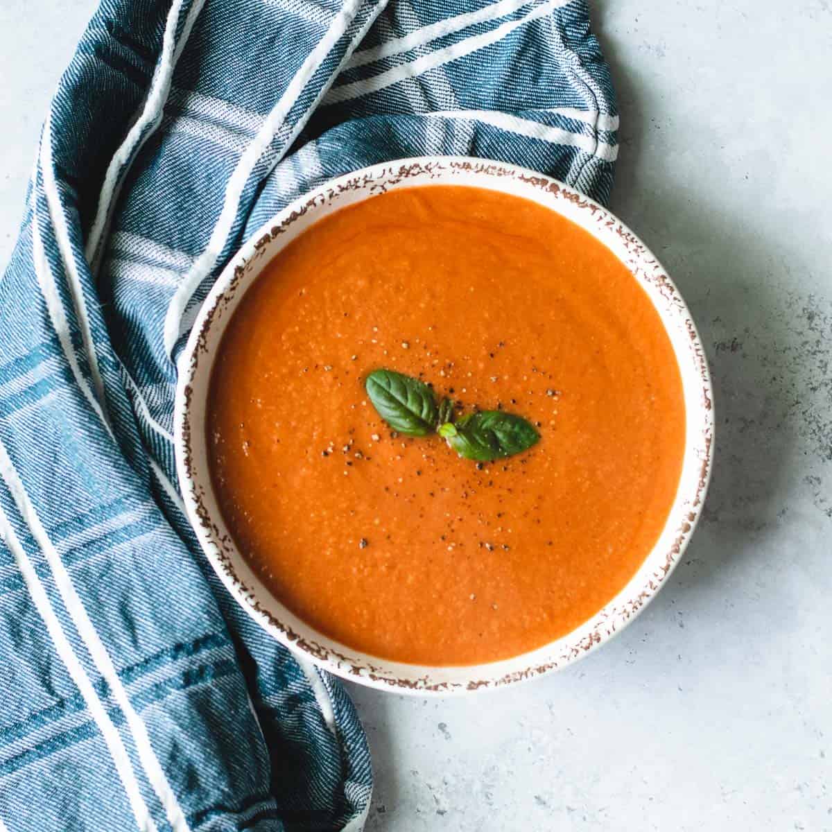 Tomato soup in a white bowl sitting on a grey background with blue and white tea towel.