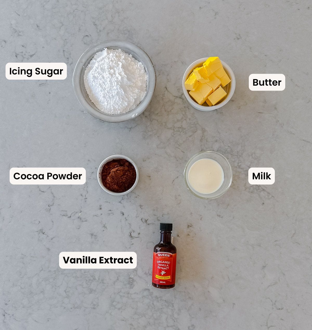 Ingredients for icing arranged on a marble surface: bowls of icing sugar, cubed butter, cocoa powder, milk, and a bottle of vanilla extract, each labelled with its name.