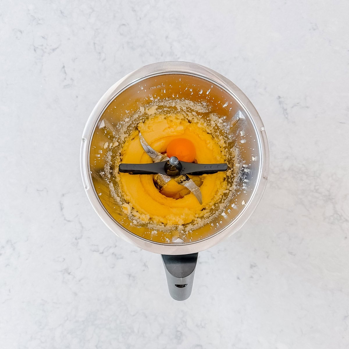 A top down view of butter and sugar creamed together in the Thermomix, with the butterfly whisk inserted and an egg yolk added.