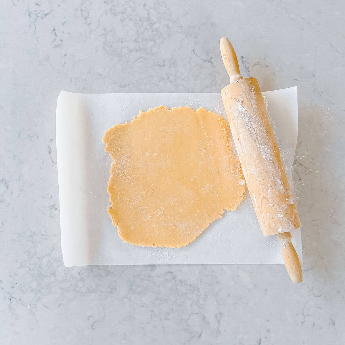 Rolled out sugar cookie dough on a sheet of baking paper, a wooden rolling pin sits on the side.
