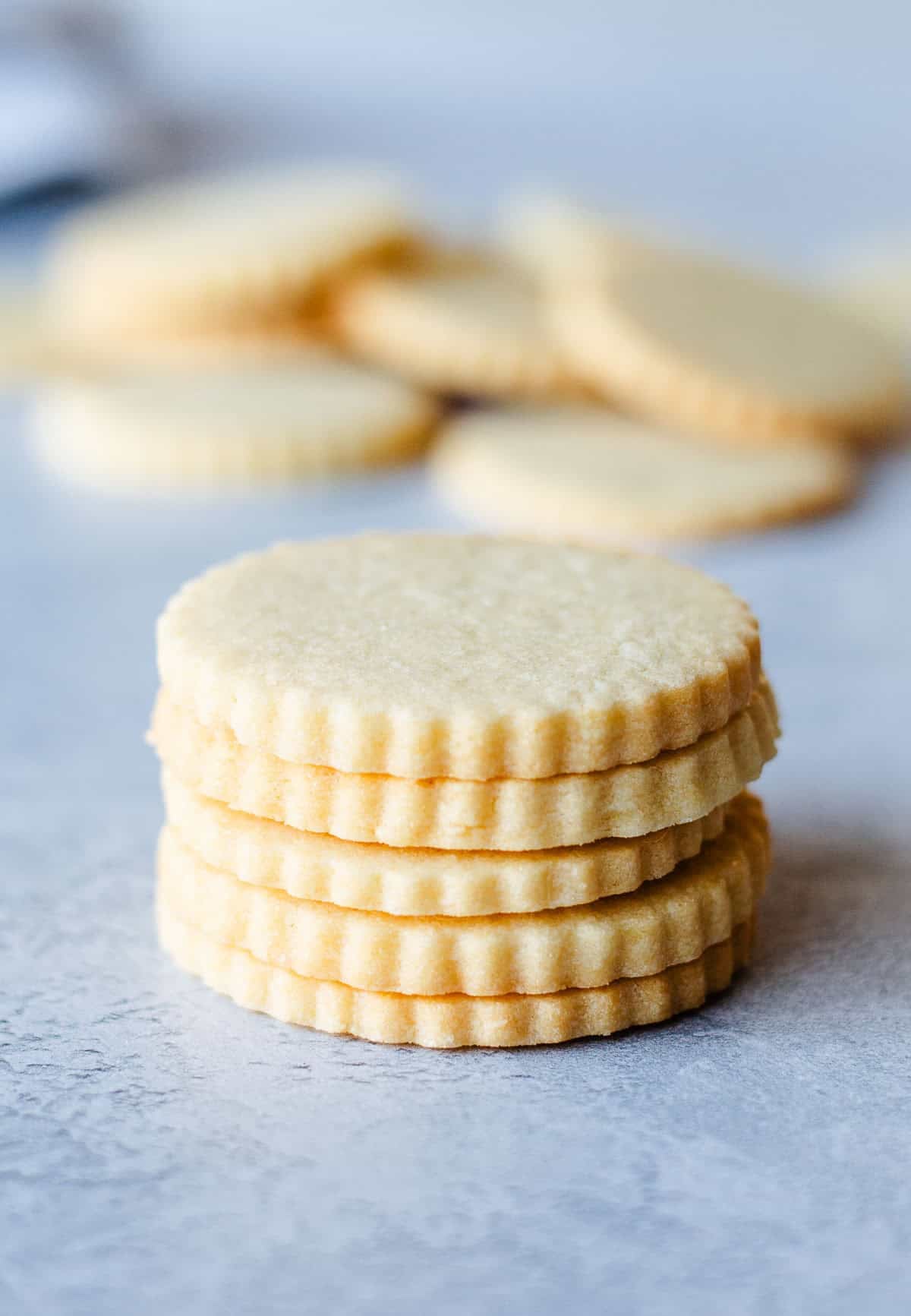 A stack of 5 sugar cookies, sitting on a grey benchtop. A handful of sugar cookies are seen in the background.