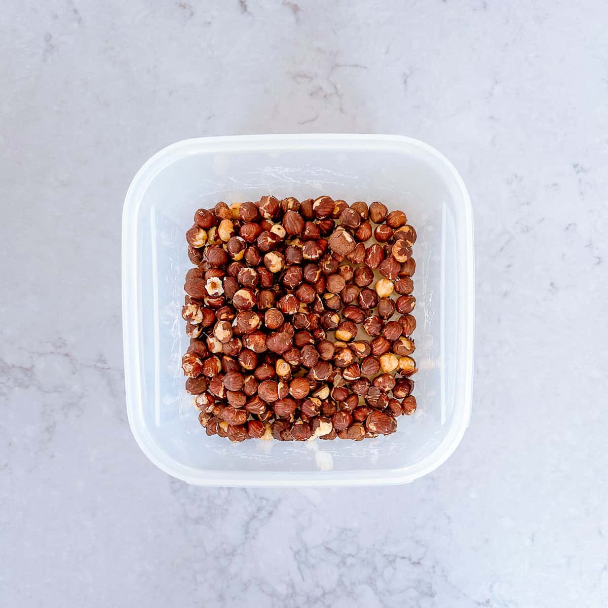 Raw hazelnuts in a plastic container on a marble benchtop.