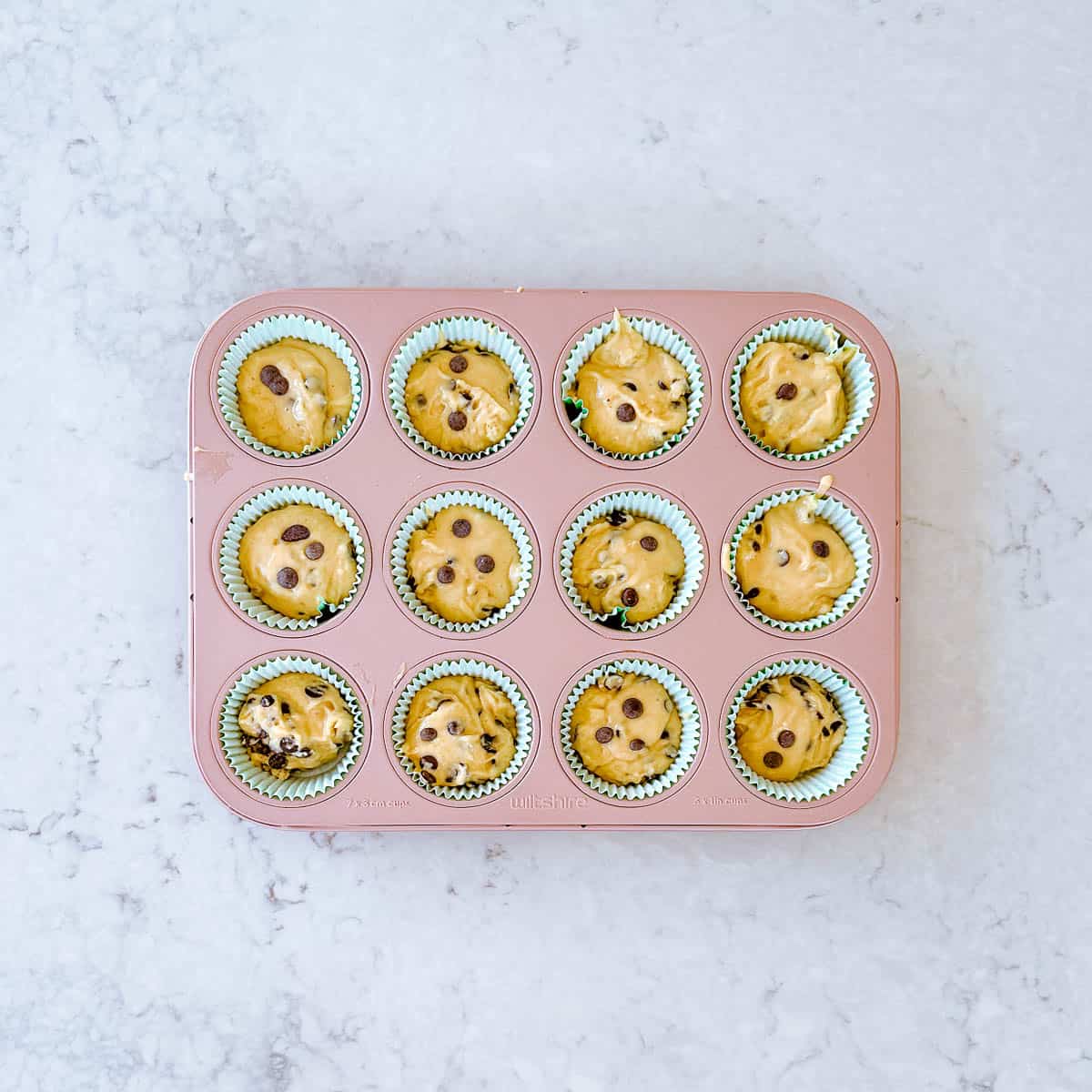 Chocolate chip muffin batter in cases in a 12 hole pink muffin tin.