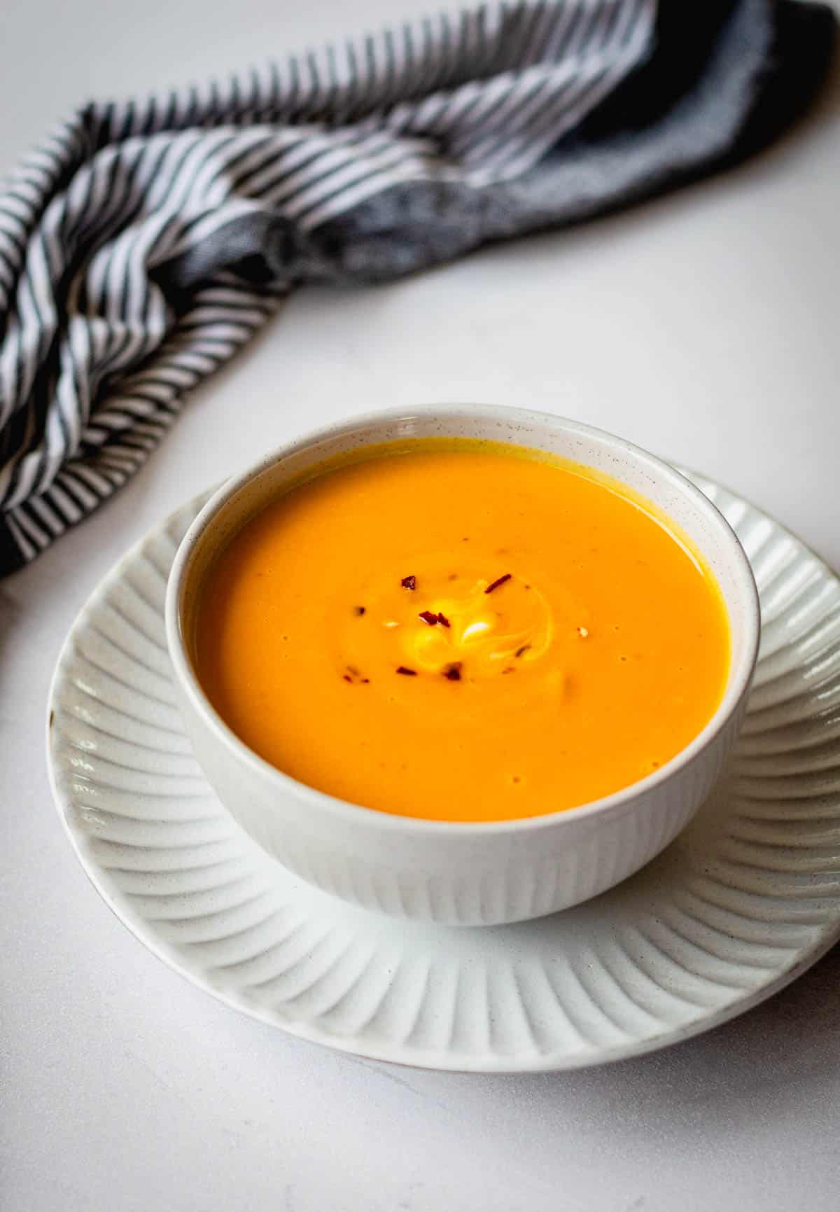 Thai Pumpkin Soup in a white bowl, sitting on a plate with a blue stripy tea towel in the background.