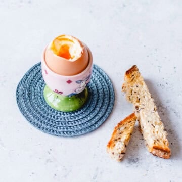 Boiled egg in a pink and green egg cup sitting on a blue coaster with toasted solders.