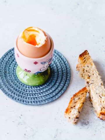 Boiled egg in a pink and green egg cup sitting on a blue coaster with toasted solders.