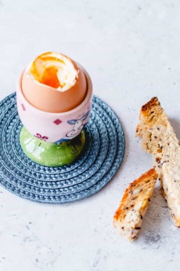 Boiled egg in a pink and green egg cup sitting on a blue coaster with toasted soldiers.