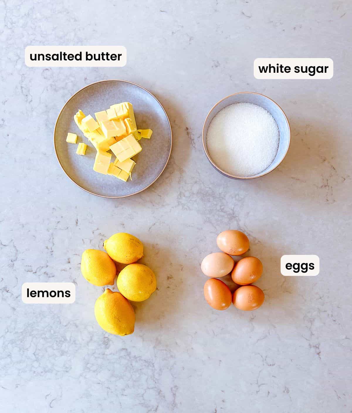 Four ingredients on a worktop: a plate of cubed unsalted butter, a bowl of white sugar, four lemons, and five brown eggs, each labelled accordingly.