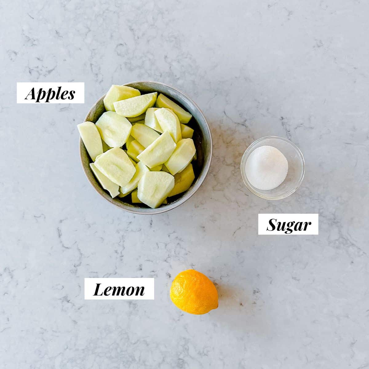 A bowl of apples, sugar and 1 lemon sitting on a marble benchtop.