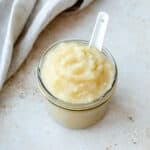 Apple sauce in a glass jar with white spoon on a stone benchtop.