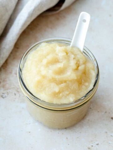 Apple sauce in a glass jar with white spoon on a stone benchtop.