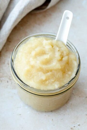 Apple sauce in a glass jar with white spoon on a stone benchtop.