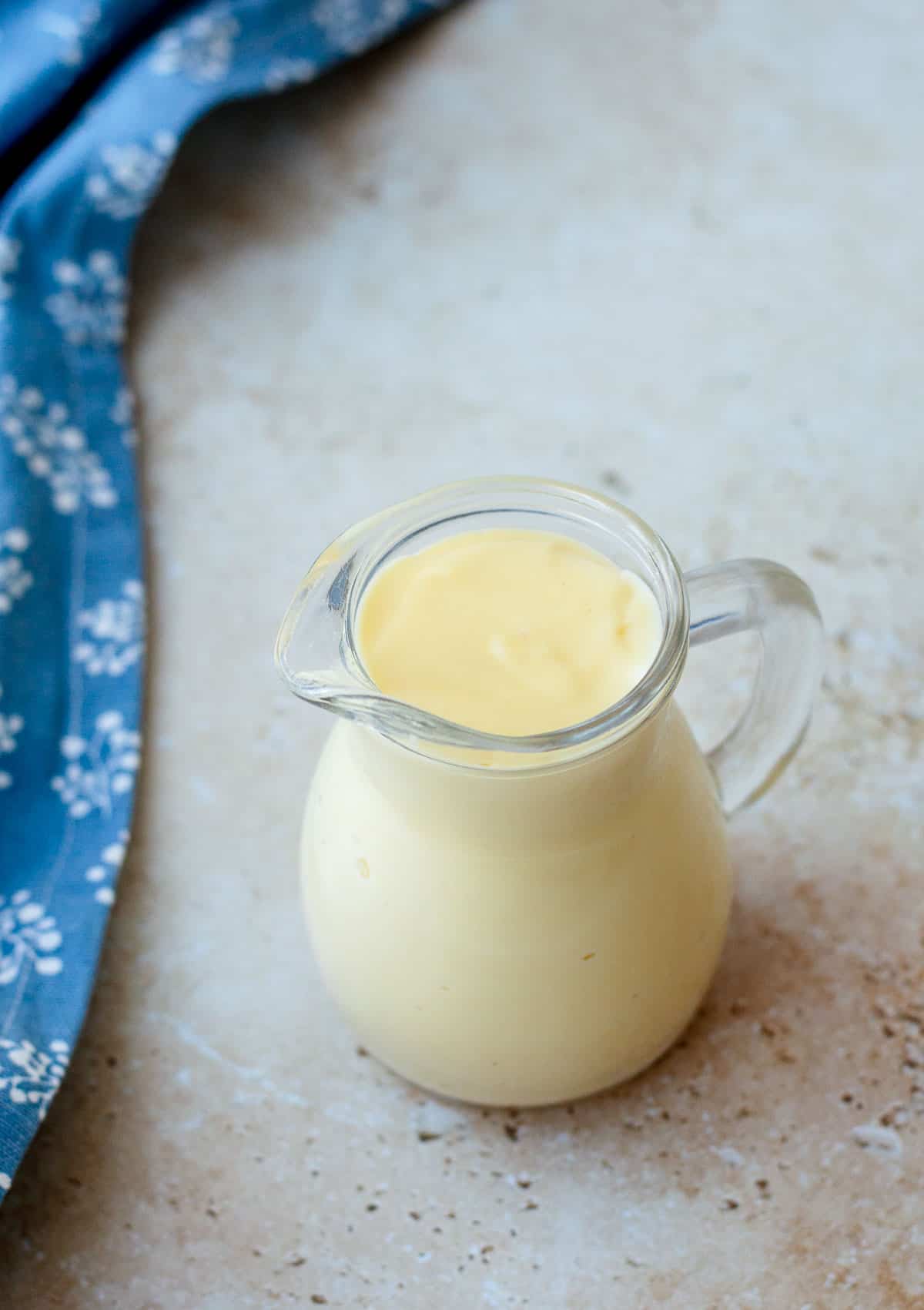 Custard in a glass jug with tea towel, sitting on a marble benchtop.