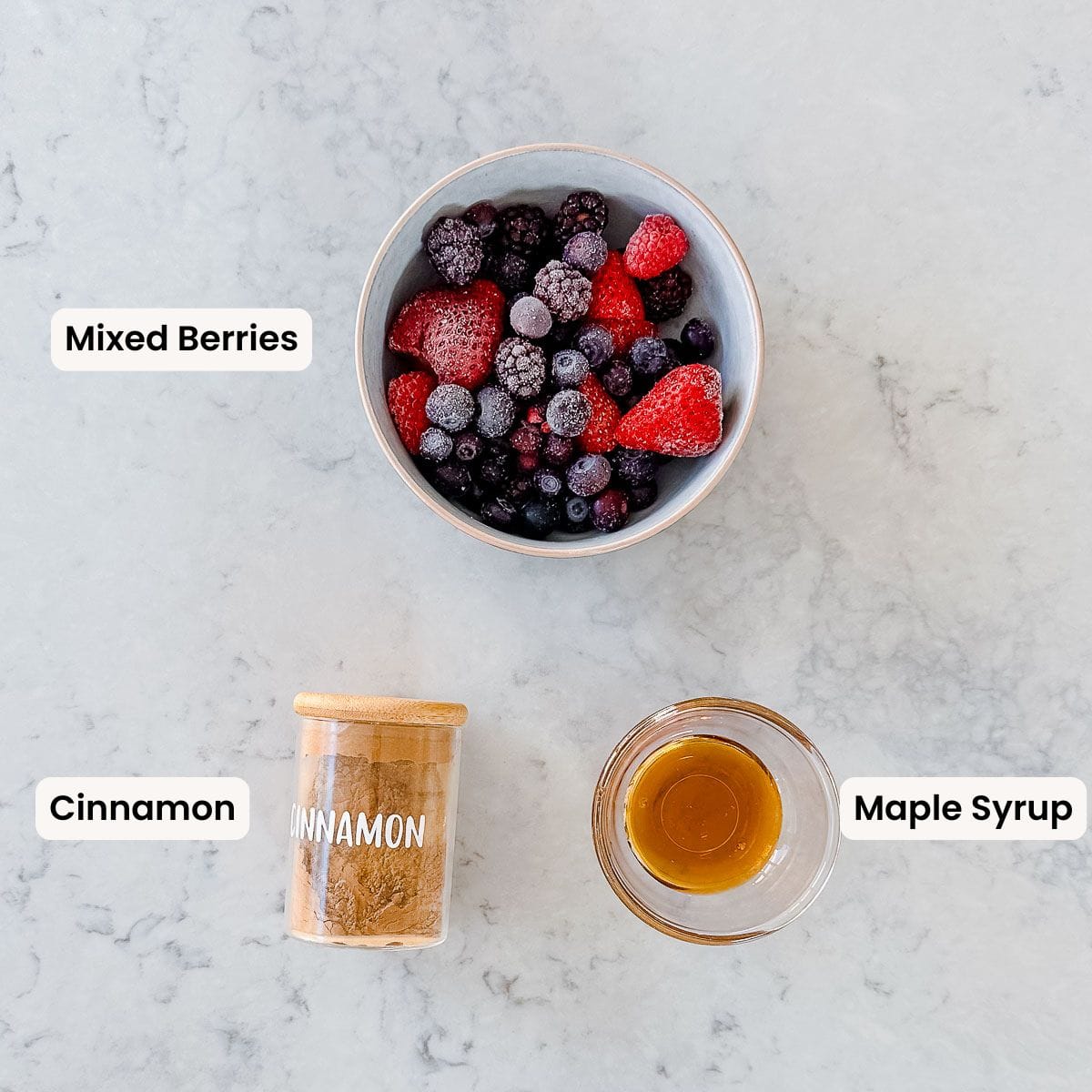 Mixed berries in a bowl, a jar of cinnamon and a small bowl of maple syrup sitting on a marble benchtop.