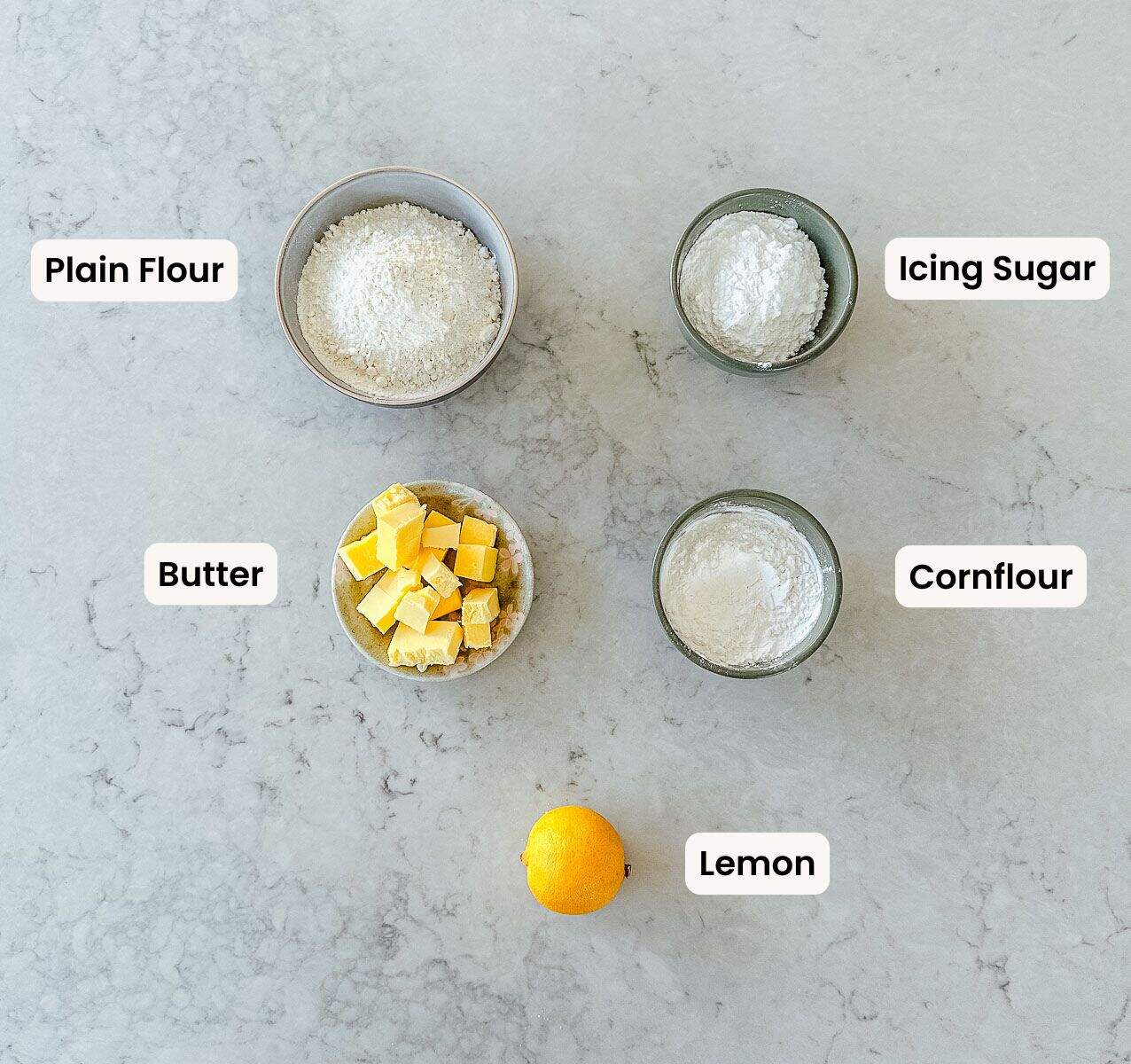 A collection of labelled ingredients used to make lemon melting moments on a marble benchtop.