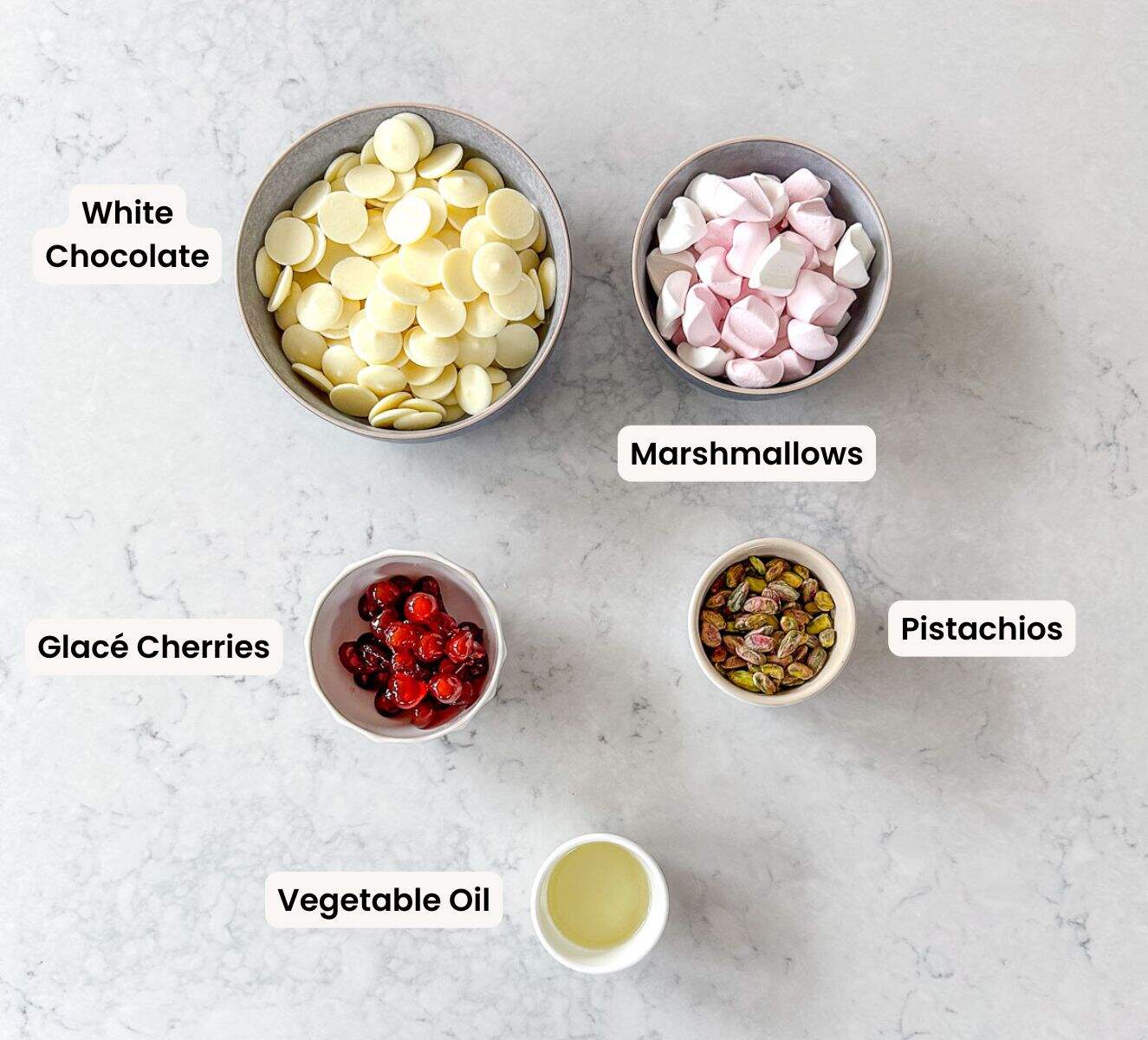 Five bowls on a light counter, containing white chocolate buttons, pink and white marshmallows, glace cherries, shelled pistachios, and vegetable oil, each labeled.
